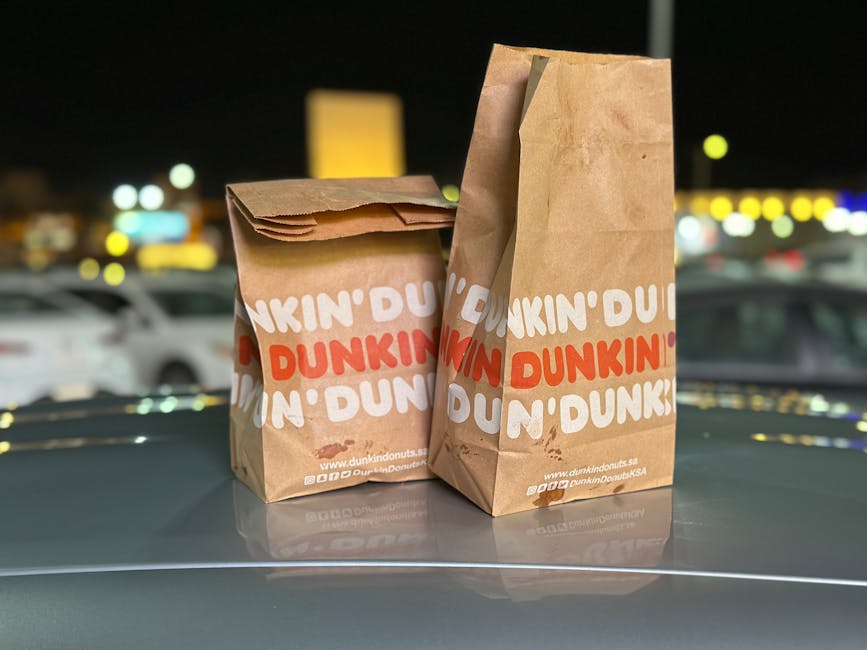 Two Dunkin' Donuts paper bags placed on a car hood with blurred night city lights in the background.