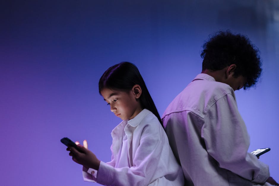 Two children using smartphones back-to-back under colorful studio lighting.