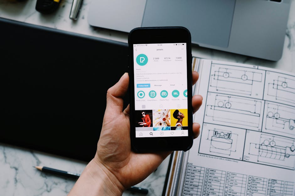Hand holding smartphone displaying social media profile with tech gadgets on desk.