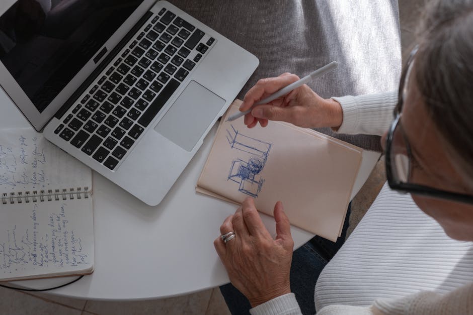 An adult woman sketching in a notebook at a desk with her laptop, working from home.